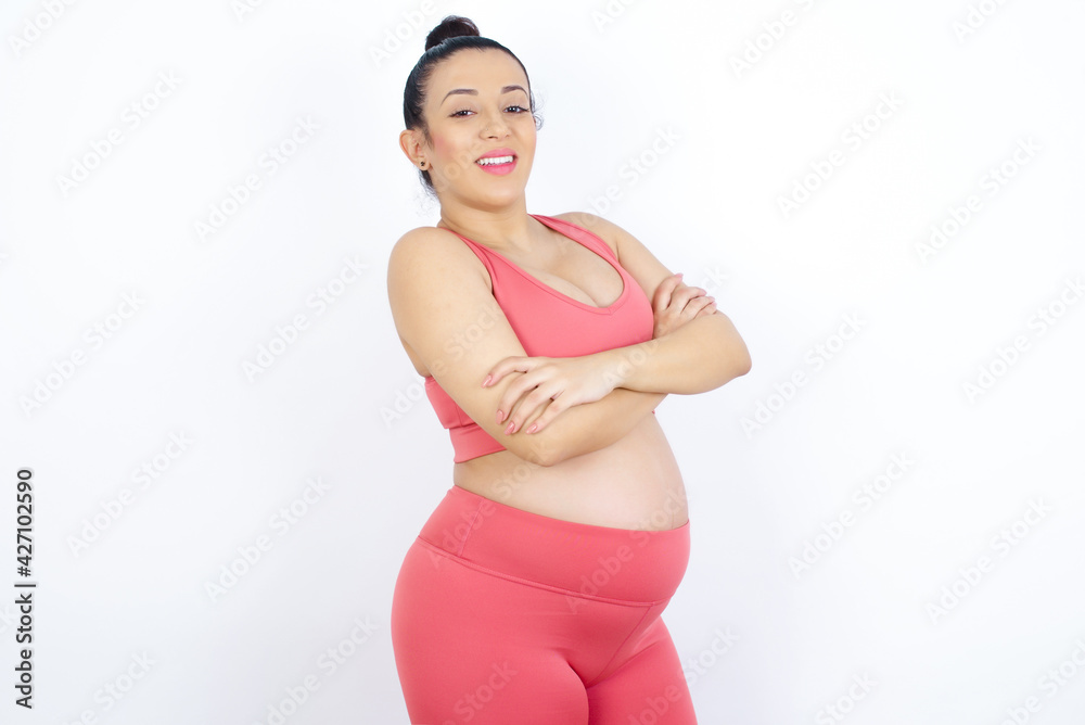 Image of cheerful young beautiful Arab pregnant woman in sports clothes against white wall with arms crossed. Looking and smiling at the camera. Confidence concept. Image of cheerful young beautiful Arab pregnant woman in sports clothes against white wall with arms crossed. Looking and smiling at the camera. Confidence concept.