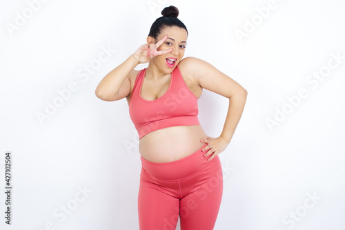 Fototapeta Naklejka Na Ścianę i Meble -  young beautiful Arab pregnant woman in sports clothes against white wall making v-sign near eyes. Leisure, coquettish, celebration, and flirt concept.