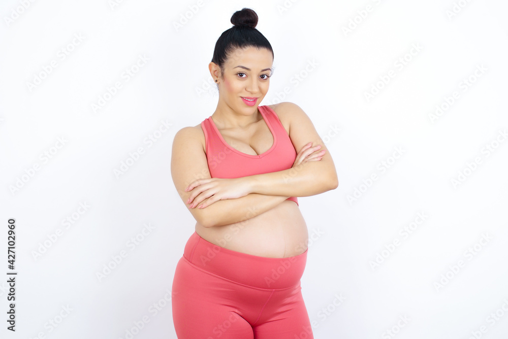 Waist up shot of self confident young beautiful Arab pregnant woman in sports clothes against white wall has broad smile, crosses arms, happy to meet with colleagues. Waist up shot of self confident young beautiful Arab pregnant woman in sports clothes against white wall has broad smile, crosses arms, happy to meet with colleagues.