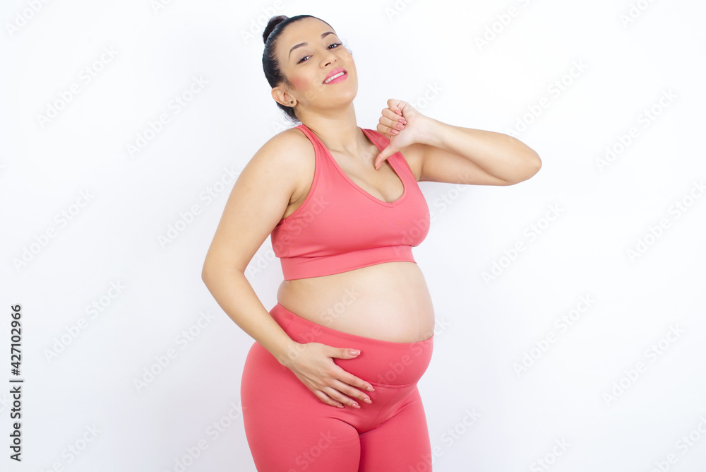 Closeup of cheerful young beautiful Arab pregnant woman in sports clothes against white wall looks joyful, satisfied and confident, points at himself with thumb. Closeup of cheerful young beautiful Arab pregnant woman in sports clothes against white wall looks joyful, satisfied and confident, points at himself with thumb.