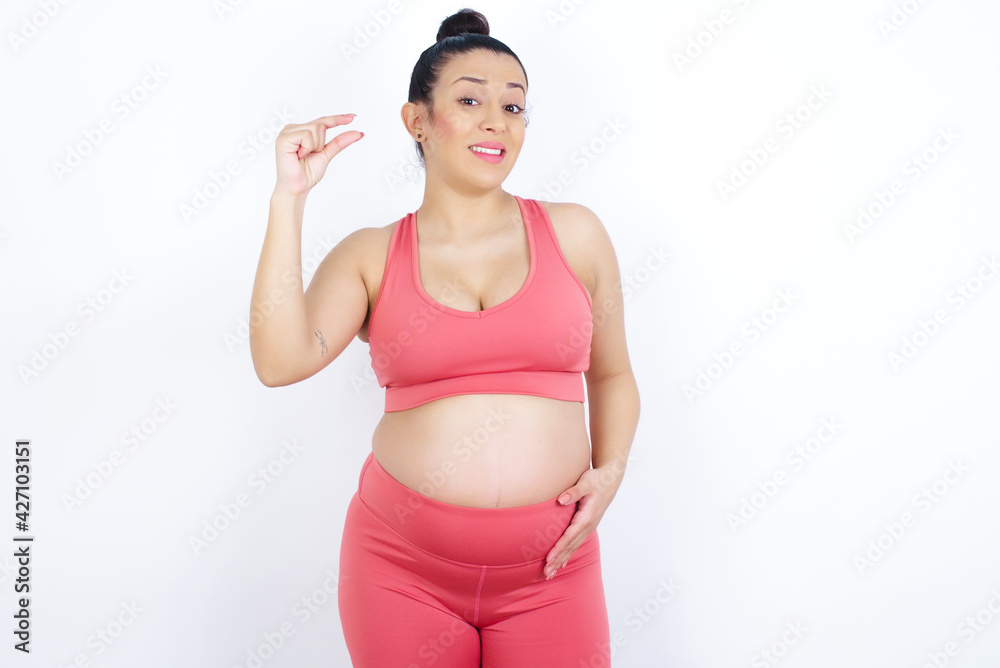 Upset young beautiful Arab pregnant woman in sports clothes against white wall shapes little gesture with hand demonstrates something very tiny small size. Not very much Upset young beautiful Arab pregnant woman in sports clothes against white wall shapes little gesture with hand demonstrates something very tiny small size. Not very much