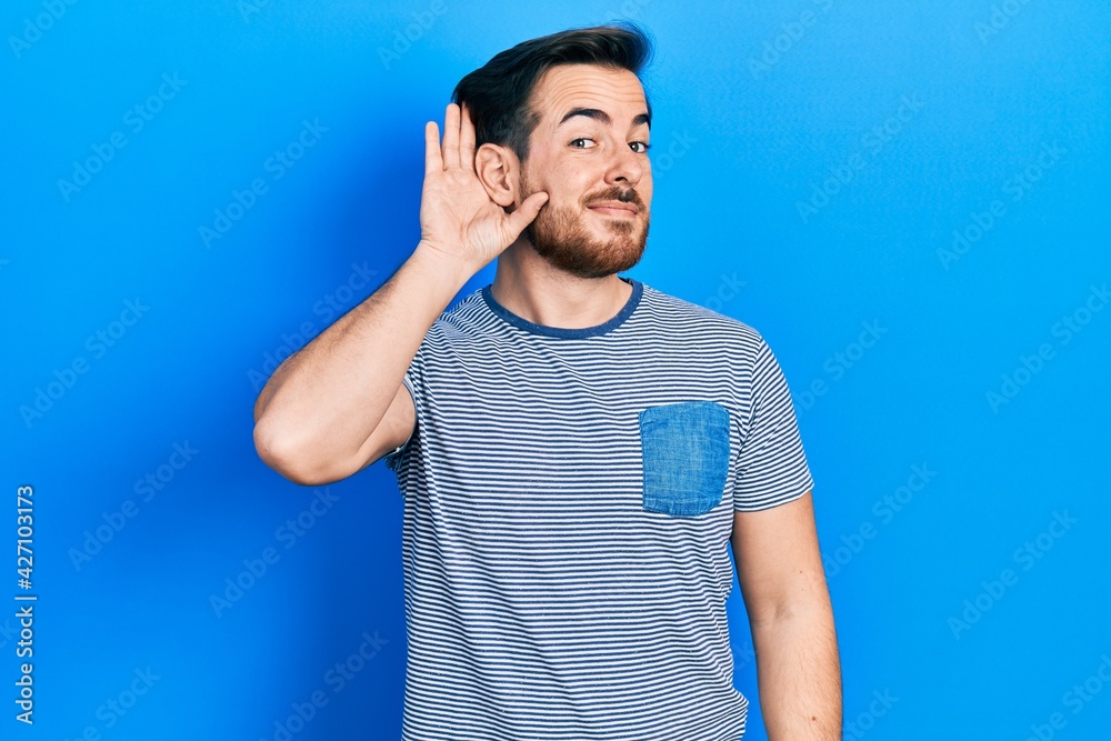 Handsome caucasian man with beard wearing casual striped t shirt smiling with hand over ear listening an hearing to rumor or gossip. deafness concept. Handsome caucasian man with beard wearing casual striped t shirt smiling with hand over ear listening an hearing to rumor or gossip. deafness concept.