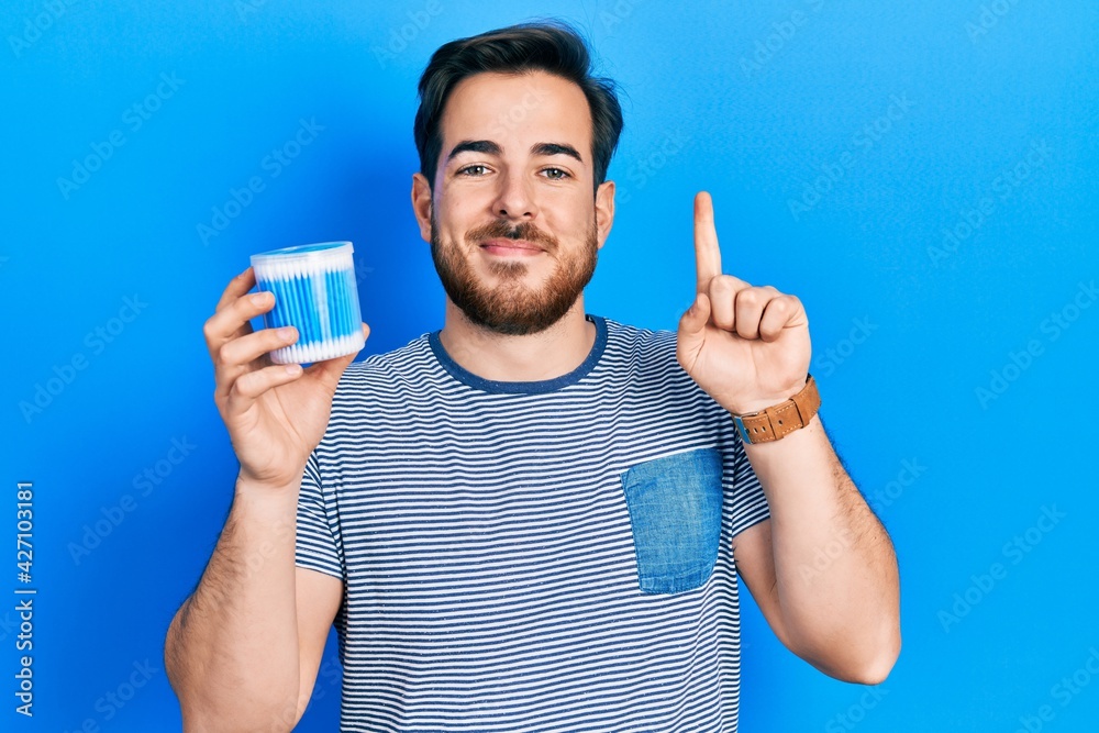 Handsome caucasian man with beard holding earwax cotton remover smiling with an idea or question pointing finger with happy face, number one Handsome caucasian man with beard holding earwax cotton remover smiling with an idea or question pointing finger with happy face, number one