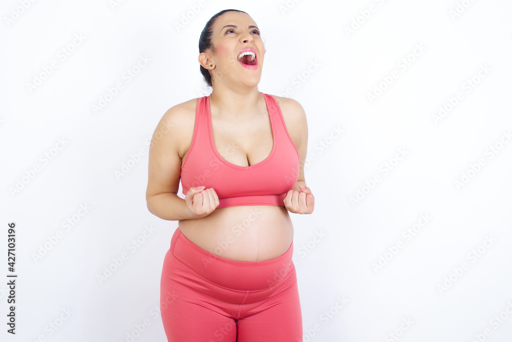 young beautiful Arab pregnant woman in sports clothes against white wall raising fists up screaming with joy being happy to achieve goals. young beautiful Arab pregnant woman in sports clothes against white wall raising fists up screaming with joy being happy to achieve goals.