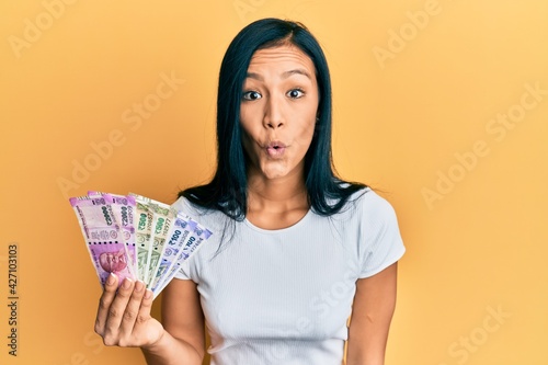 Beautiful hispanic woman holding indian rupee banknotes scared and amazed with open mouth for surprise, disbelief face