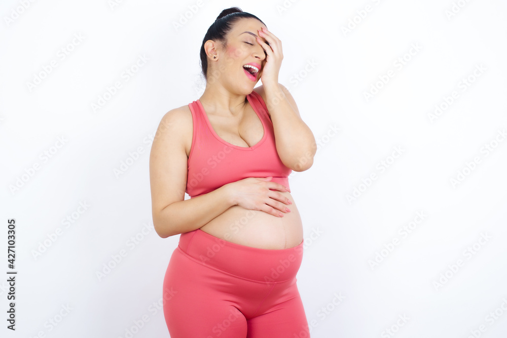 Charismatic carefree joyful young beautiful Arab pregnant woman in sports clothes against white wall likes laugh out loud not hiding emotions giggling hear funny hilarious joke chuckling facepalm. Charismatic carefree joyful young beautiful Arab pregnant woman in sports clothes against white wall likes laugh out loud not hiding emotions giggling hear funny hilarious joke chuckling facepalm.