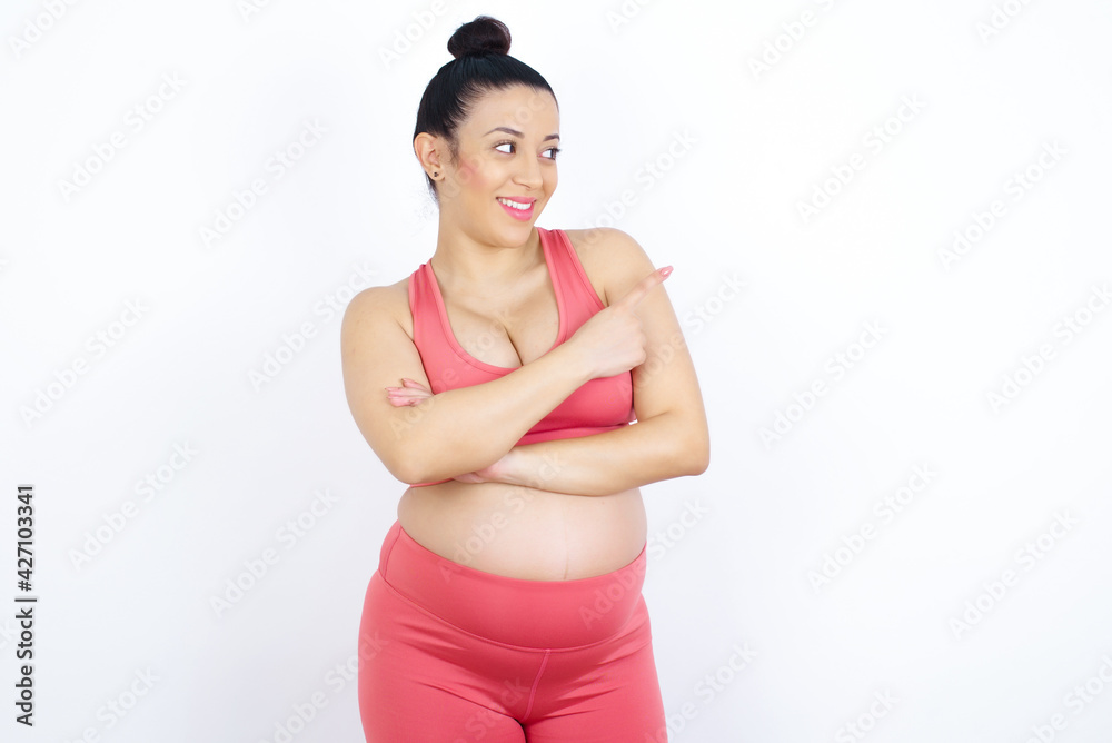 Happy cheerful smiling young beautiful Arab pregnant woman in sports clothes against white wall looking and pointing aside with hand. Copy space and advertisement concept. Happy cheerful smiling young beautiful Arab pregnant woman in sports clothes against white wall looking and pointing aside with hand. Copy space and advertisement concept.