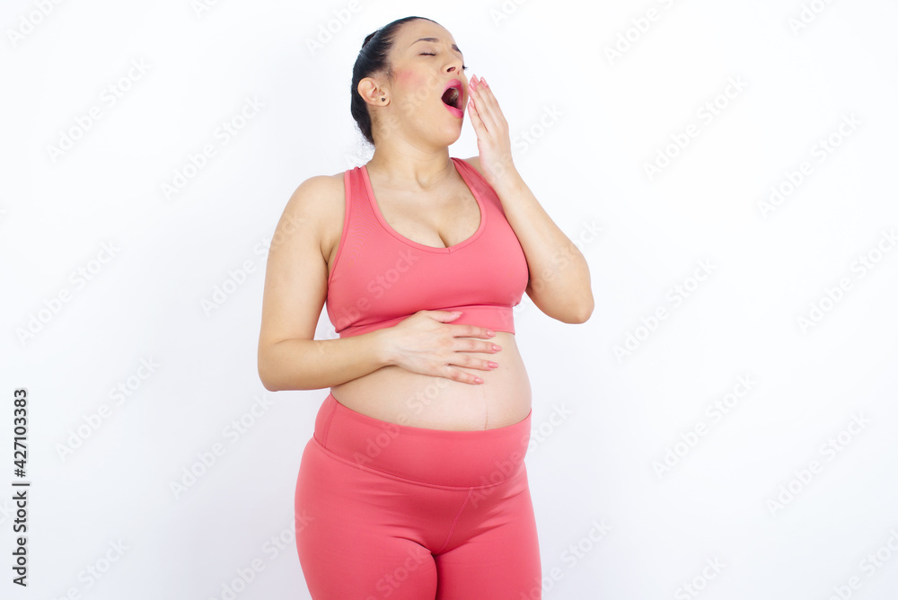 Sleepy young beautiful Arab pregnant woman in sports clothes against white wall yawning with messy hair, feeling tired after sleepless night, yawning, covering mouth with palm. Sleepy young beautiful Arab pregnant woman in sports clothes against white wall yawning with messy hair, feeling tired after sleepless night, yawning, covering mouth with palm.