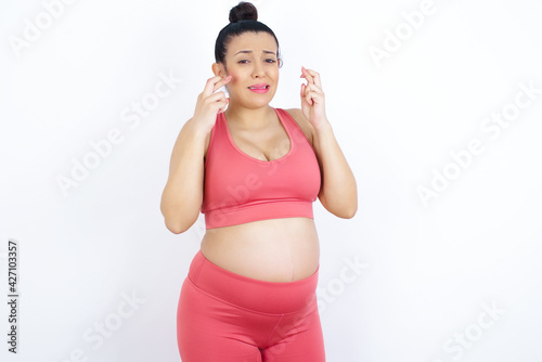 young beautiful Arab pregnant woman in sports clothes against white wall holding fingers crossed with worried expression hoping boss didn't noticed mistakes at work. © Jihan