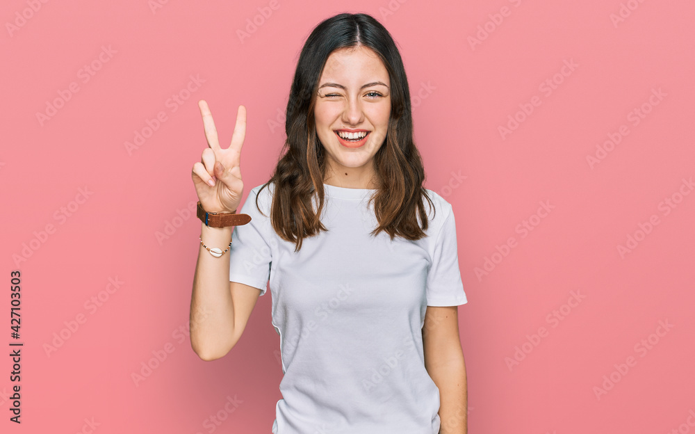 Young beautiful woman wearing casual white t shirt smiling with happy face winking at the camera doing victory sign with fingers. number two. Young beautiful woman wearing casual white t shirt smiling with happy face winking at the camera doing victory sign with fingers. number two.