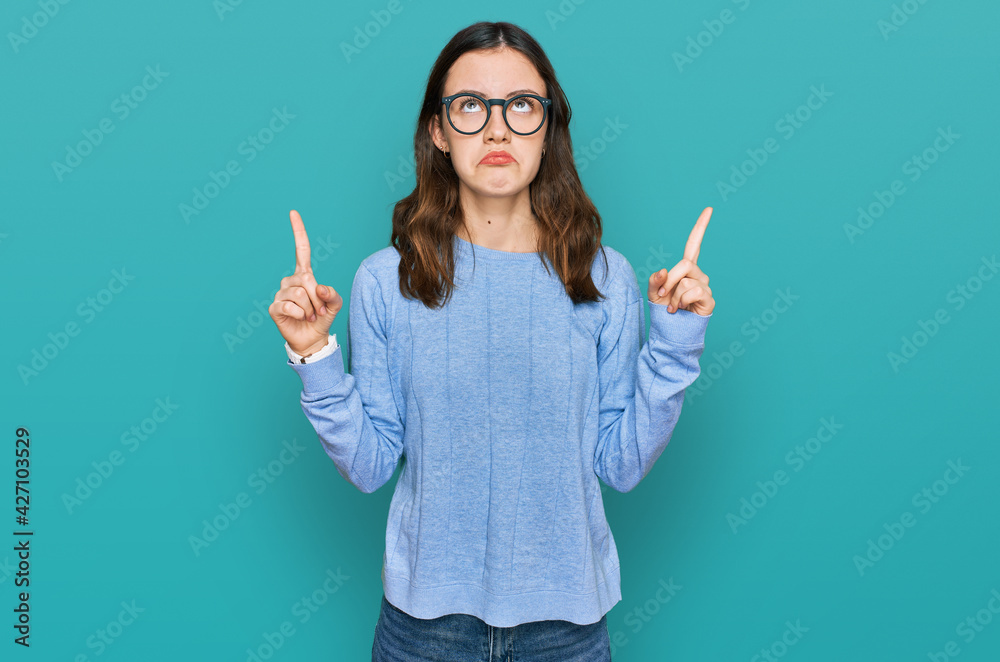 Young beautiful woman wearing casual clothes and glasses pointing up looking sad and upset, indicating direction with fingers, unhappy and depressed. Young beautiful woman wearing casual clothes and glasses pointing up looking sad and upset, indicating direction with fingers, unhappy and depressed.