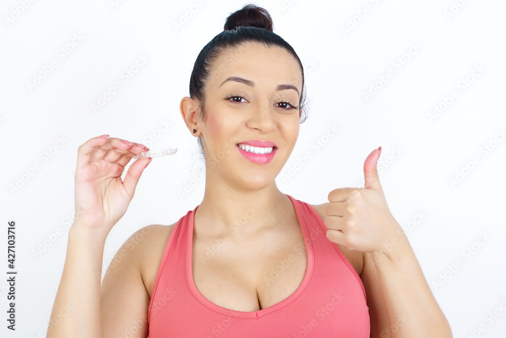 young beautiful Arab woman in sports clothes against white wall holding an invisible braces aligner and rising thumb up, recommending this new treatment. Dental healthcare concept. young beautiful Arab woman in sports clothes against white wall holding an invisible braces aligner and rising thumb up, recommending this new treatment. Dental healthcare concept.