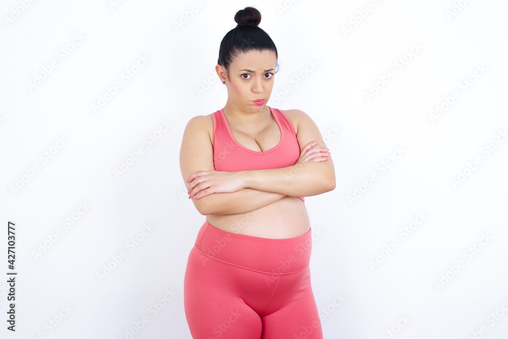 Gloomy dissatisfied young beautiful Arab pregnant woman in sports clothes against white wall looks with miserable expression at camera from under forehead, makes unhappy grimace Gloomy dissatisfied young beautiful Arab pregnant woman in sports clothes against white wall looks with miserable expression at camera from under forehead, makes unhappy grimace