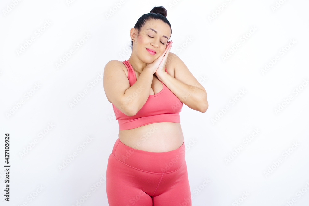 young beautiful Arab pregnant woman in sports clothes against white wall leans on pressed palms closes eyes and has pleasant smile dreams about something young beautiful Arab pregnant woman in sports clothes against white wall leans on pressed palms closes eyes and has pleasant smile dreams about something