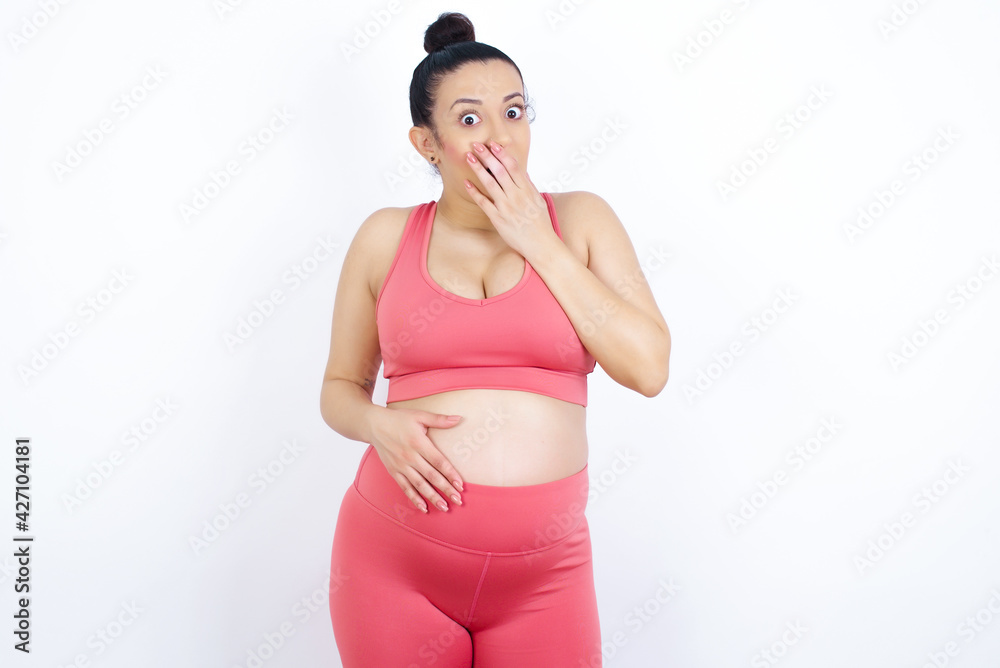 young beautiful Arab pregnant woman in sports clothes against white wall covers mouth and looks with wonder at camera, cannot believe unexpected rumors.