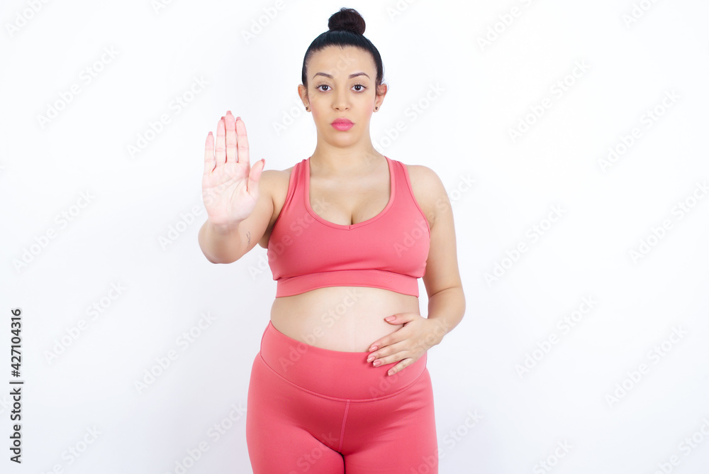 young beautiful Arab pregnant woman in sports clothes against white wall shows stop sign prohibition symbol keeps palm forward to camera with strict expression young beautiful Arab pregnant woman in sports clothes against white wall shows stop sign prohibition symbol keeps palm forward to camera with strict expression