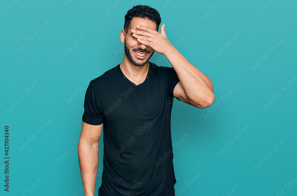 Hispanic man with beard wearing casual black t shirt smiling and laughing with hand on face covering eyes for surprise. blind concept. Hispanic man with beard wearing casual black t shirt smiling and laughing with hand on face covering eyes for surprise. blind concept.