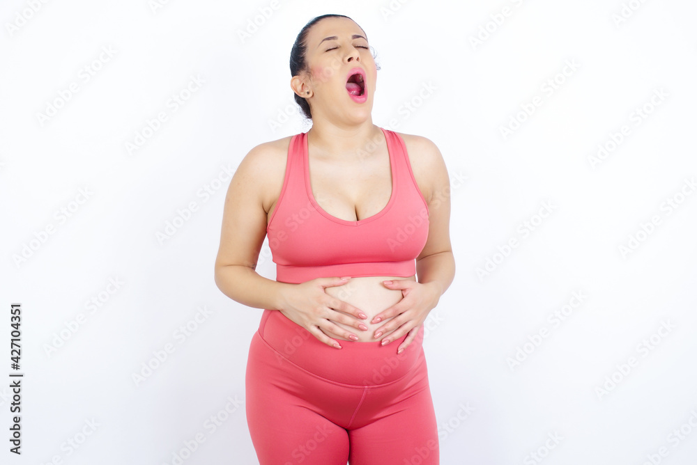 young beautiful Arab pregnant woman in sports clothes against white wall yawns with opened mouth stands. Daily morning routine young beautiful Arab pregnant woman in sports clothes against white wall yawns with opened mouth stands. Daily morning routine