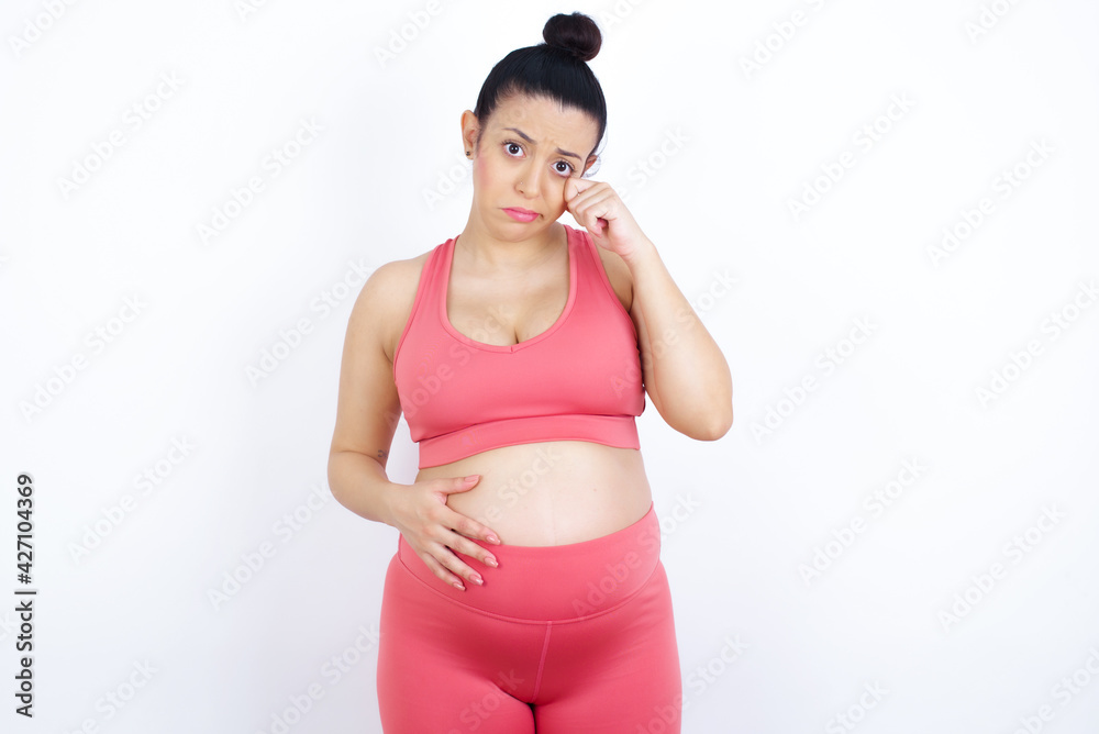 Disappointed dejected young beautiful Arab pregnant woman in sports clothes against white wall wipes tears stands stressed with gloomy expression. Negative emotion Disappointed dejected young beautiful Arab pregnant woman in sports clothes against white wall wipes tears stands stressed with gloomy expression. Negative emotion