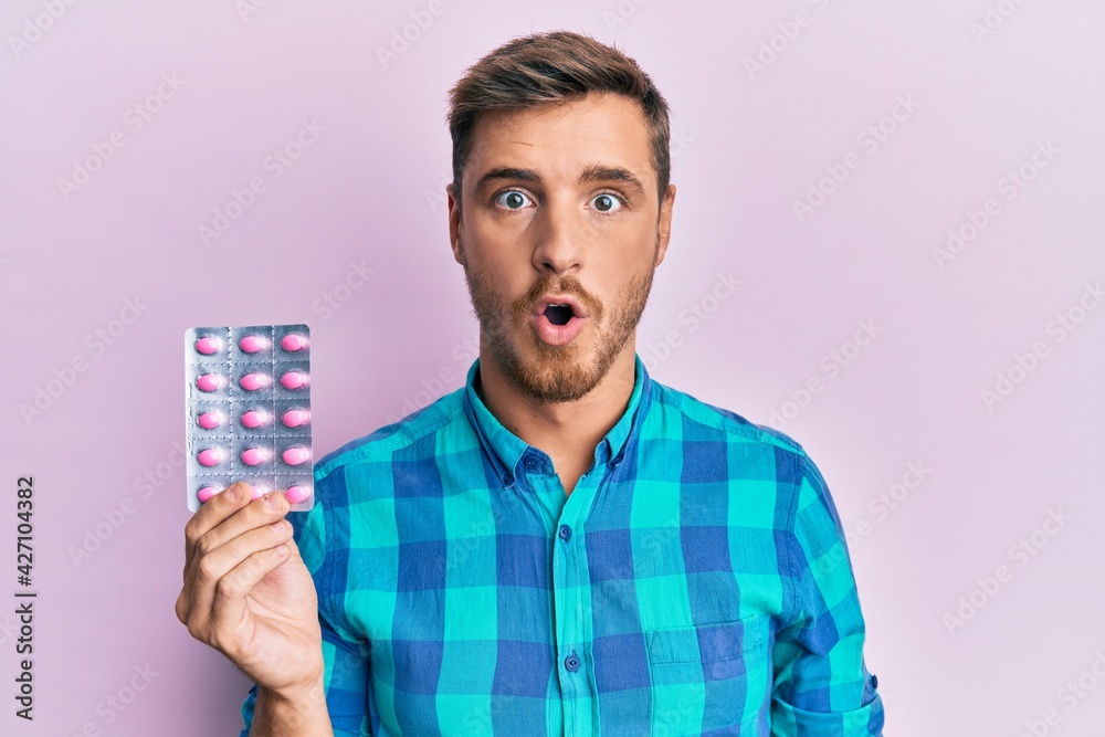 Handsome caucasian man holding pills scared and amazed with open mouth for surprise, disbelief face Handsome caucasian man holding pills scared and amazed with open mouth for surprise, disbelief face
