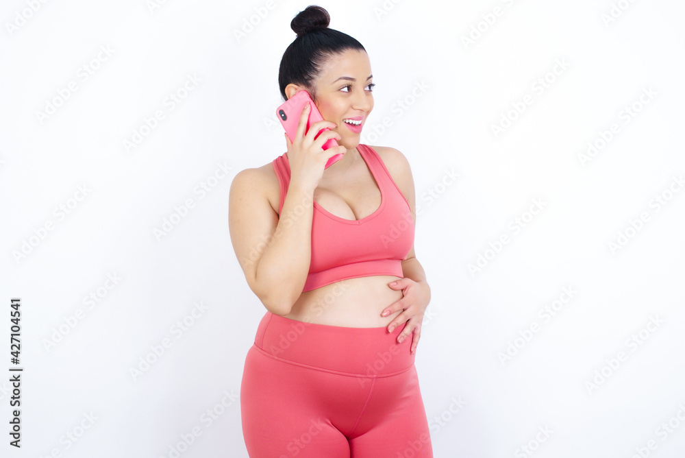 Pleasant looking happy young beautiful Arab pregnant woman in sports clothes has nice telephone conversation and looks aside, has nice mood and smiles positively while talks via cell phone Pleasant looking happy young beautiful Arab pregnant woman in sports clothes has nice telephone conversation and looks aside, has nice mood and smiles positively while talks via cell phone