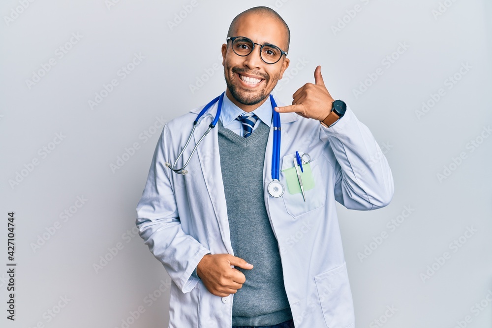 Hispanic adult man wearing doctor uniform and stethoscope smiling doing phone gesture with hand and fingers like talking on the telephone. communicating concepts. Hispanic adult man wearing doctor uniform and stethoscope smiling doing phone gesture with hand and fingers like talking on the telephone. communicating concepts.