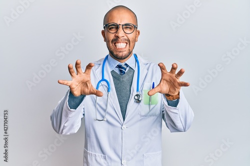 Hispanic adult man wearing doctor uniform and stethoscope smiling funny doing claw gesture as cat, aggressive and sexy expression