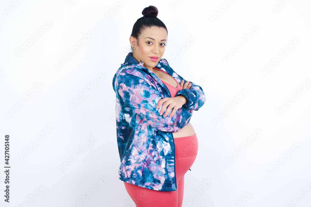 Portrait of young beautiful Arab pregnant woman in sports clothes against white wall standing with folded arms and smiling
Portrait of young beautiful Arab pregnant woman in sports clothes against white wall standing with folded arms and smiling
