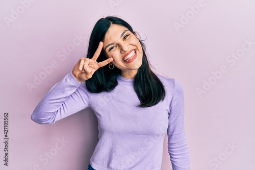 Young hispanic woman wearing casual clothes doing peace symbol with fingers over face, smiling cheerful showing victory