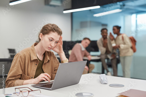 Portrait of stressed young woman using laptop in office or coworking space with group of people in background, copy space