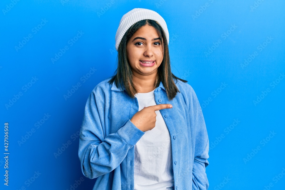 Young latin woman wearing cute wool cap pointing aside worried and nervous with forefinger, concerned and surprised expression Young latin woman wearing cute wool cap pointing aside worried and nervous with forefinger, concerned and surprised expression