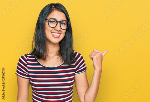 Fototapeta Naklejka Na Ścianę i Meble -  Beautiful asian young woman wearing casual clothes and glasses smiling with happy face looking and pointing to the side with thumb up.