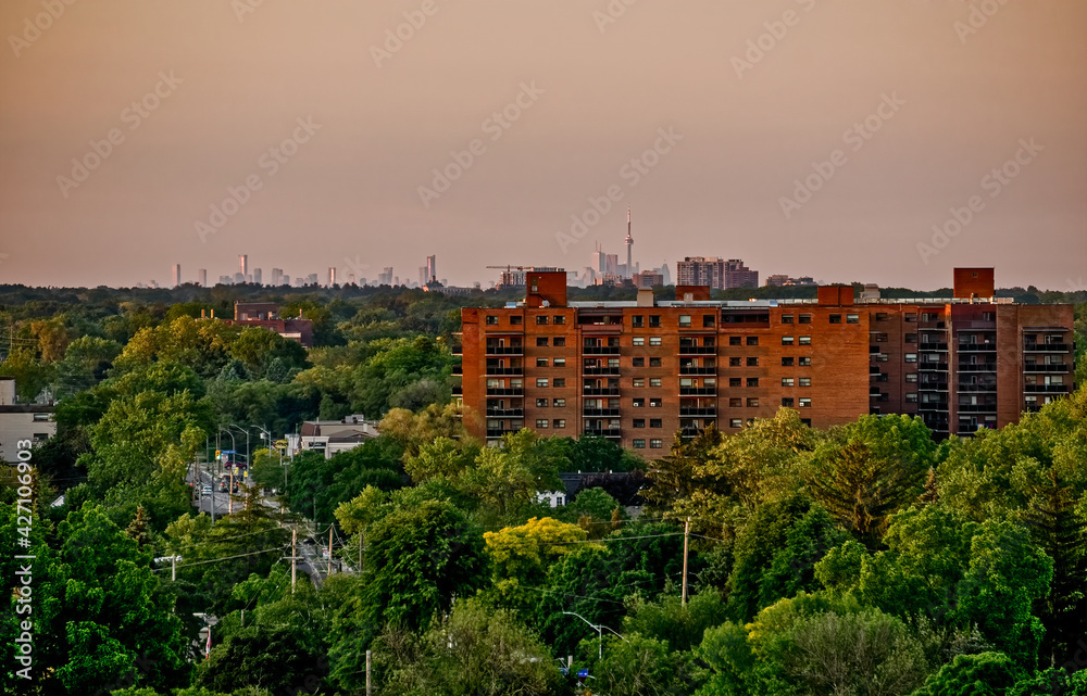 The Toronto skyline is seen in the distance behind an apartment building set in a green urban forest at sunset in the summer