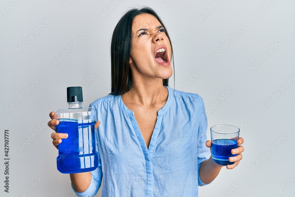 Young hispanic girl holding mouthwash for fresh breath angry and mad screaming frustrated and furious, shouting with anger looking up. Young hispanic girl holding mouthwash for fresh breath angry and mad screaming frustrated and furious, shouting with anger looking up.
