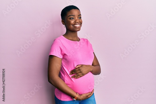 Young african american woman expecting a baby, touching pregnant belly looking away to side with smile on face, natural expression. laughing confident.