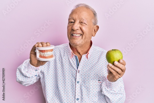 Senior caucasian man holding denture and green apple winking looking at the camera with sexy expression, cheerful and happy face. photo