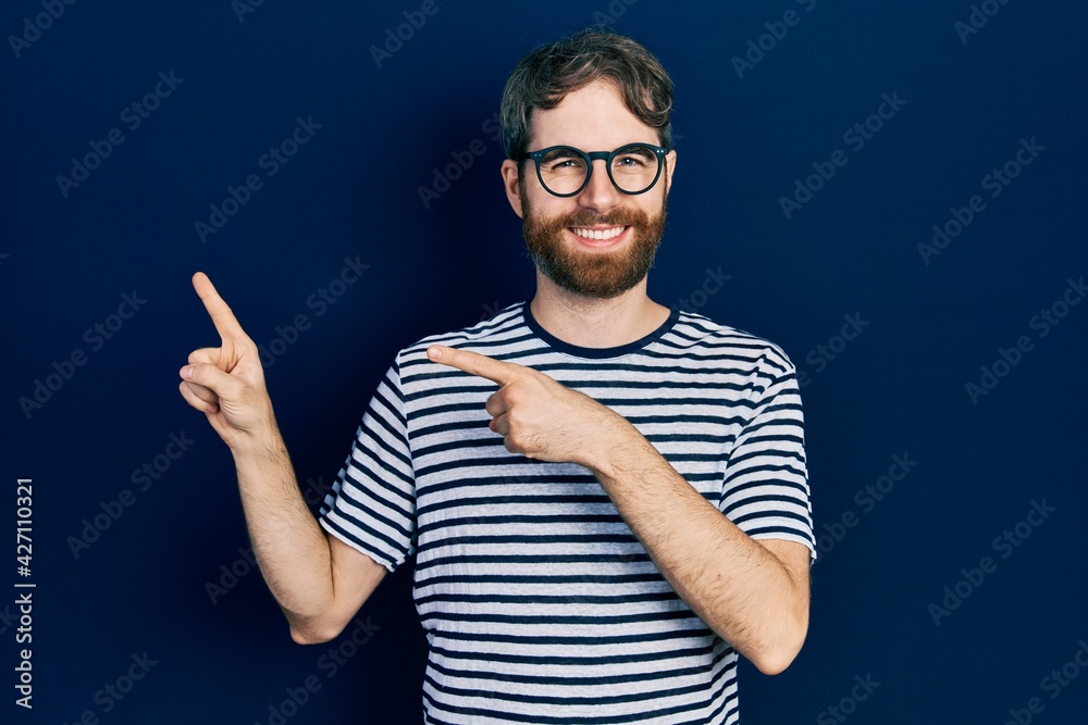 Caucasian man with beard wearing striped t shirt and glasses smiling and looking at the camera pointing with two hands and fingers to the side. Caucasian man with beard wearing striped t shirt and glasses smiling and looking at the camera pointing with two hands and fingers to the side.
