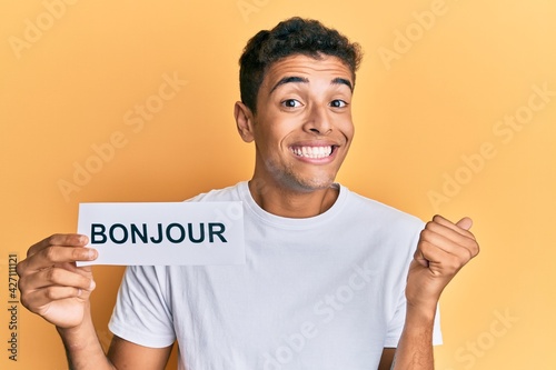 Young handsome african american man holding bonjour french greeting word screaming proud, celebrating victory and success very excited with raised arm © Krakenimages.com