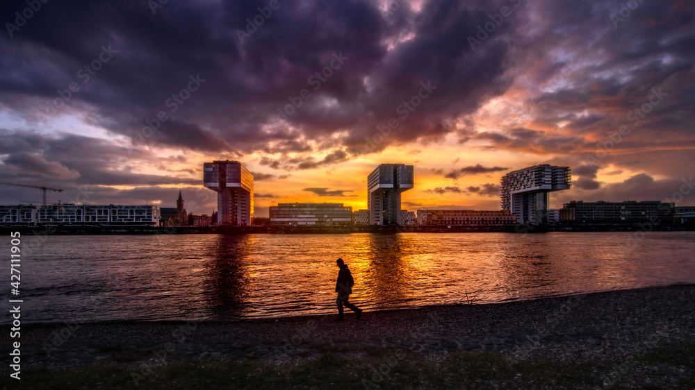 The image was shot in the beautiful city of Köln with the gorgeous Kranhäuser im Rheinauhafen and a magnificent golden hour sky and a sillouhette of an unknown passer by in the foreground.