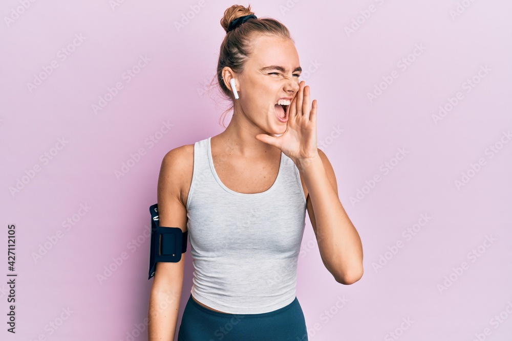 Beautiful blonde sport woman wearing arm band and earphones shouting and screaming loud to side with hand on mouth. communication concept.