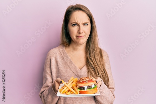 Young blonde woman eating a tasty classic burger with fries skeptic and nervous, frowning upset because of problem. negative person.