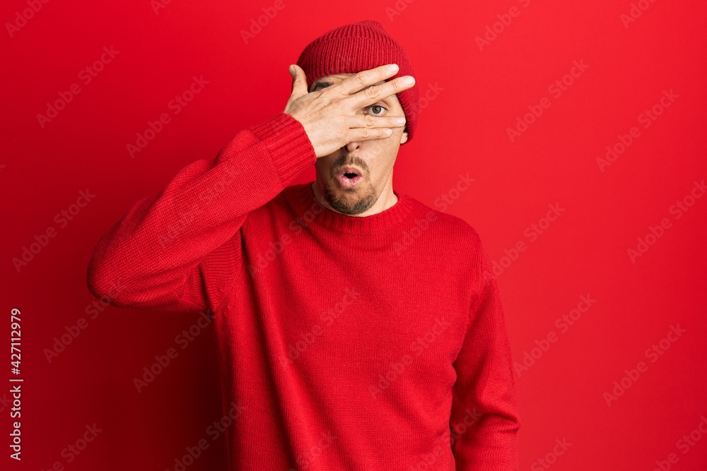 Bald man with beard wearing wool sweater and winter hat peeking in shock covering face and eyes with hand, looking through fingers afraid Bald man with beard wearing wool sweater and winter hat peeking in shock covering face and eyes with hand, looking through fingers afraid