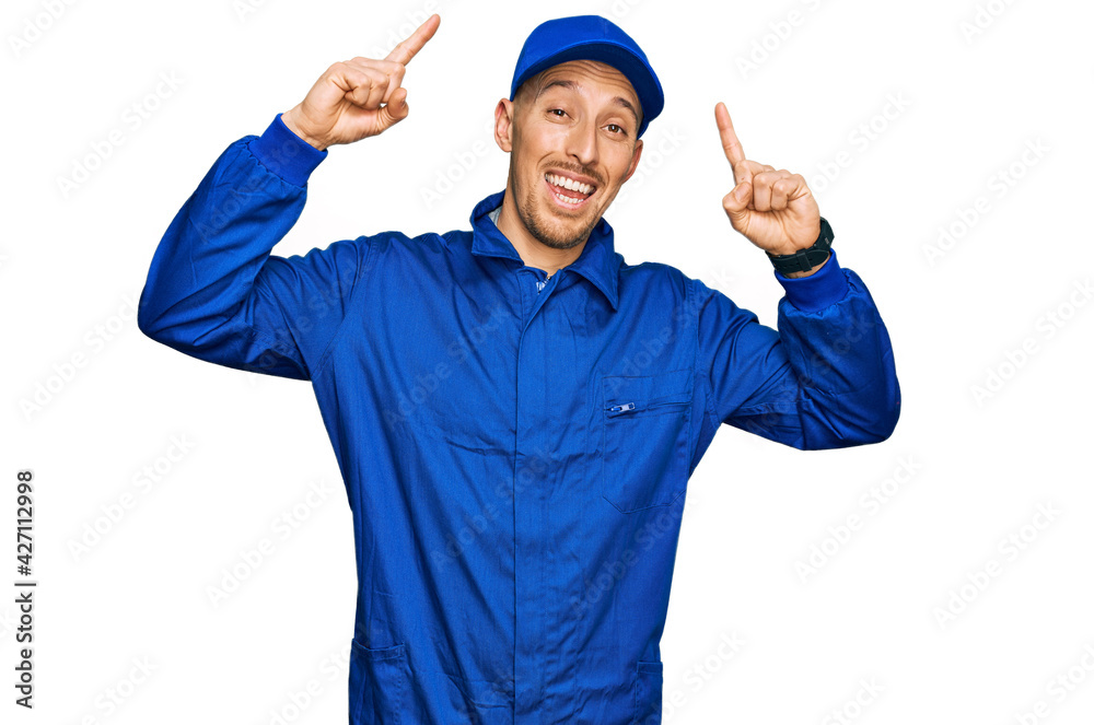 Bald man with beard wearing builder jumpsuit uniform smiling amazed and surprised and pointing up with fingers and raised arms.