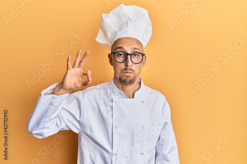 Bald man with beard wearing professional cook apron doing ok gesture puffing cheeks with funny face. mouth inflated with air, catching air.