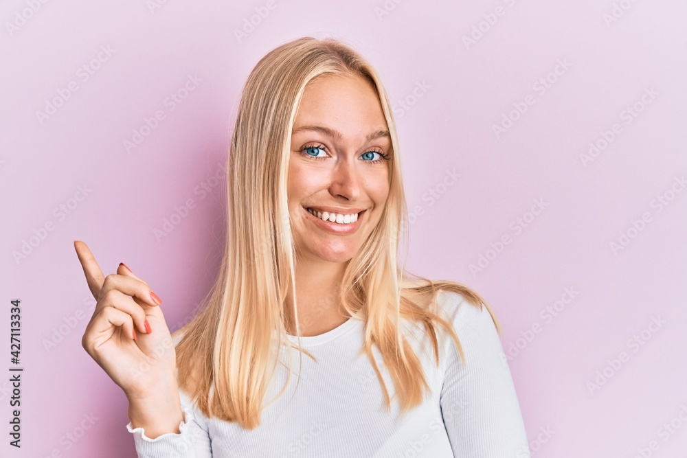 Young blonde girl wearing casual clothes with a big smile on face, pointing with hand and finger to the side looking at the camera.