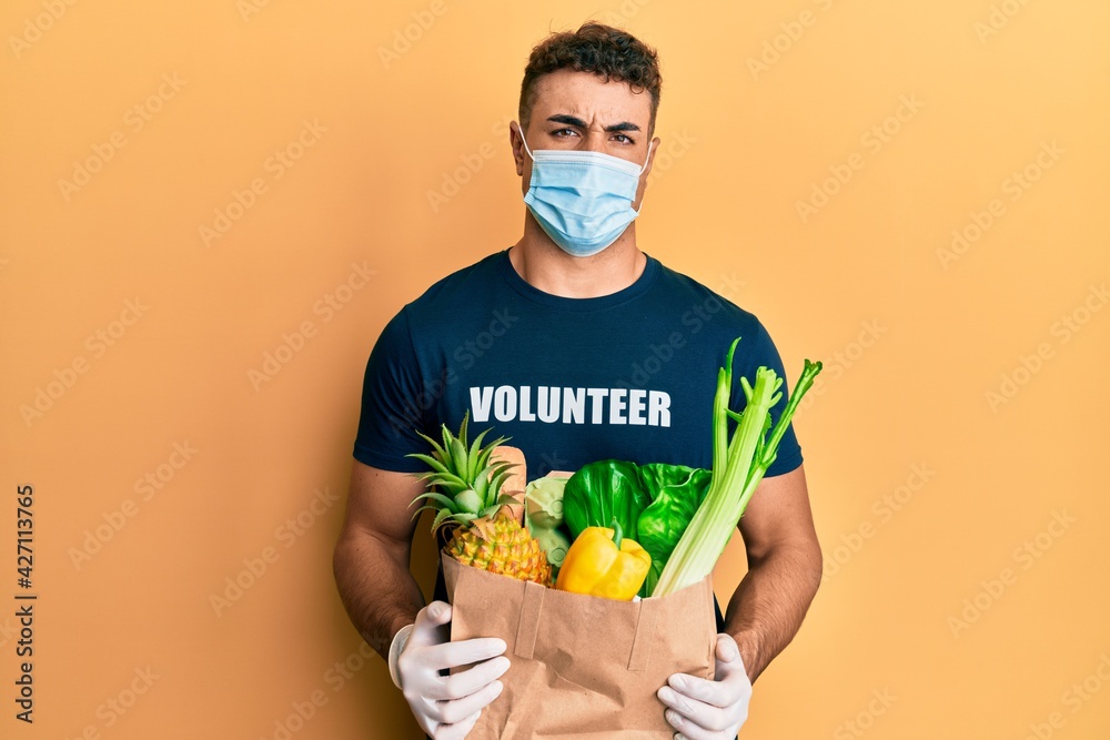 Hispanic young man wearing medical mask holding paper bag with groceries clueless and confused expression. doubt concept. Hispanic young man wearing medical mask holding paper bag with groceries clueless and confused expression. doubt concept.