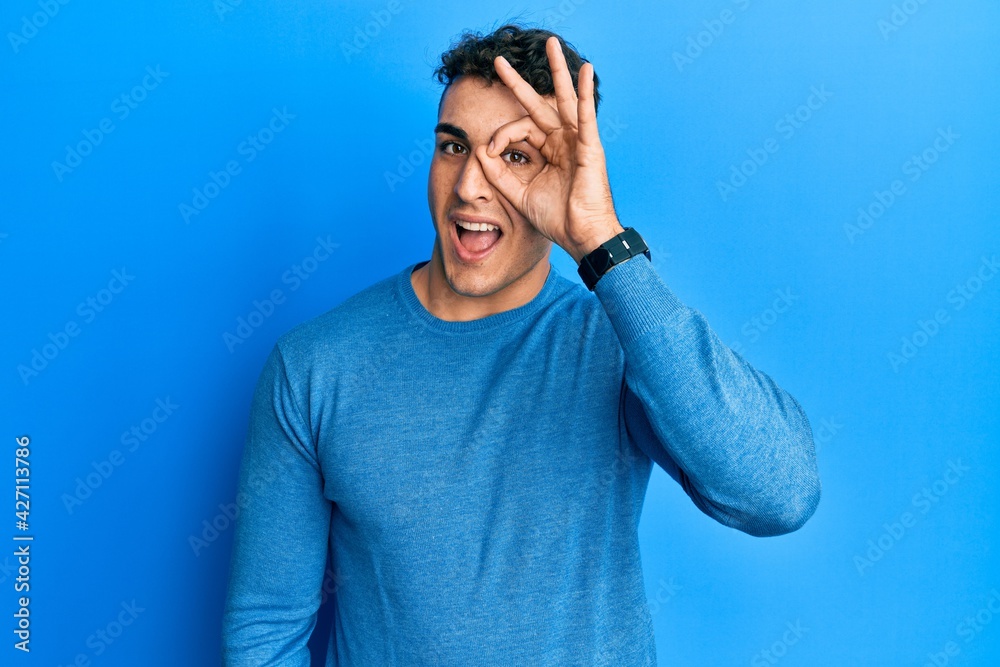 Hispanic young man wearing casual winter sweater doing ok gesture with hand smiling, eye looking through fingers with happy face. Hispanic young man wearing casual winter sweater doing ok gesture with hand smiling, eye looking through fingers with happy face.