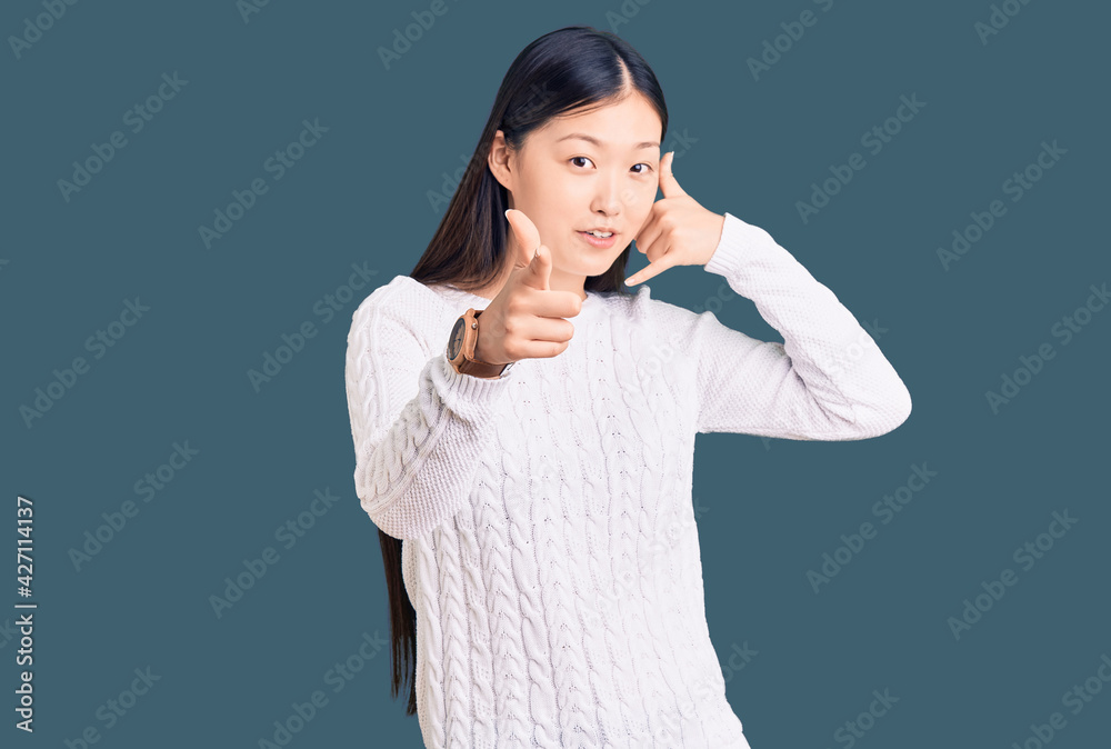 Young beautiful chinese woman wearing casual sweater smiling doing talking on the telephone gesture and pointing to you. call me. Young beautiful chinese woman wearing casual sweater smiling doing talking on the telephone gesture and pointing to you. call me.