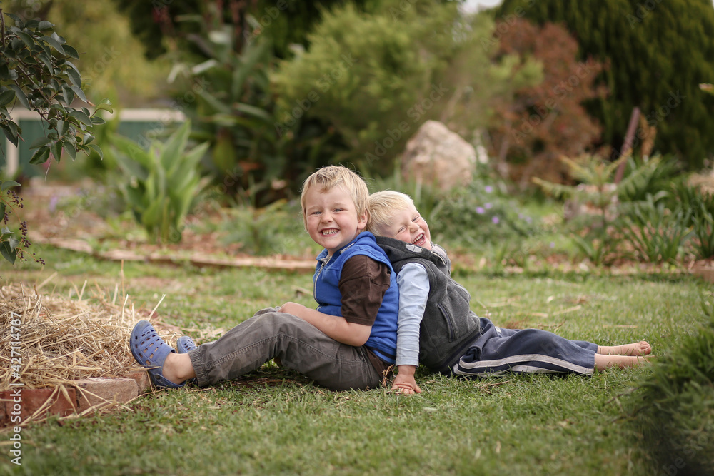 Beautiful blonde brothers sitting back to back on grass in cute garden with happy smiles Beautiful blonde brothers sitting back to back on grass in cute garden with happy smiles