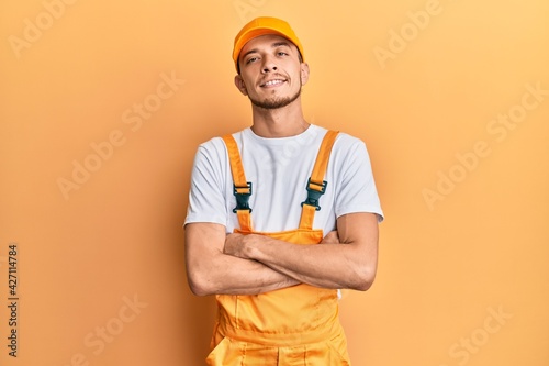Hispanic young man wearing handyman uniform happy face smiling with crossed arms looking at the camera. positive person.